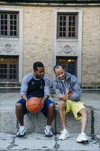 Black men sitting on concrete with basketball and texting on cell phone