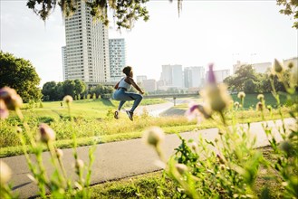 Mixed Race woman jumping on running path in park beyond wildflowers