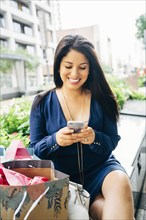 Hispanic woman with shopping bags sitting on bench texting on cell phone
