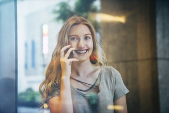 Smiling Caucasian woman talking on cell phone behind window