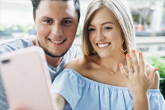 Caucasian couple posing for cell phone selfie showing engagement ring