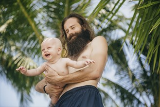 Caucasian father holding baby daughter under palm tree
