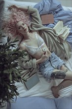 Caucasian woman with pink hair laying on bed holding flower