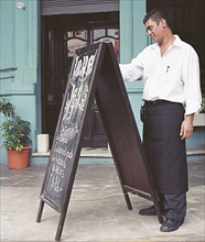Hispanic waiter writing on chalkboard in front of restaurant