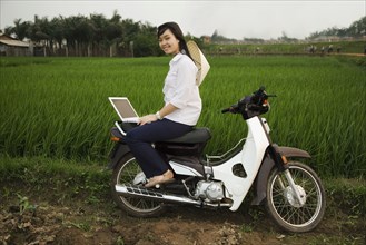 Asian woman sitting on motor scooter with laptop