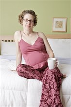 Caucasian pregnant woman drinking cup of coffee on bed