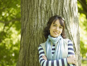 Smiling Hispanic woman leaning against tree with arms crossed