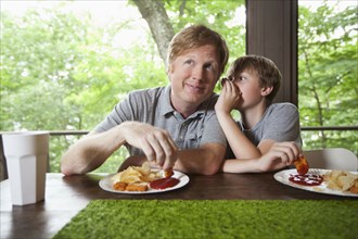 Caucasian son whispering to father and dipping food in ketchup