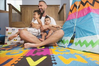 Father sitting on floor of playroom holding son and daughter