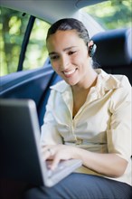 Hispanic businesswoman working in rear seat of car with laptop