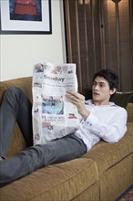 Mixed race man laying on sofa reading newspaper