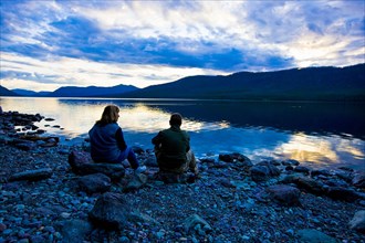 Caucasian couple sitting on rocks at lake during sunset