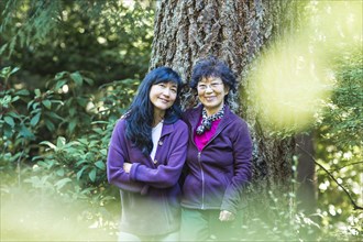 Smiling Japanese mother and daughter posing near tree trunk