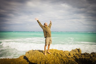 Caucasian man standing on rocks at ocean with arms raised