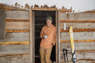 Mixed race man drinking coffee in log cabin doorway in snow