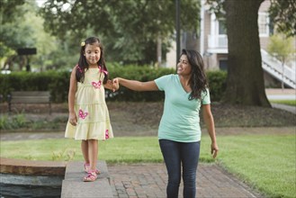 Hispanic mother holding hand of daughter walking on park wall