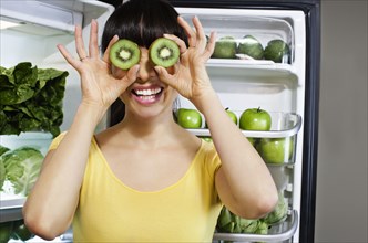 Mixed race woman covering eyes with kiwi slices near refrigerator