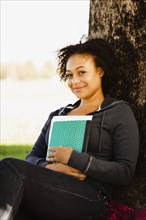 African woman holding book leaning against tree