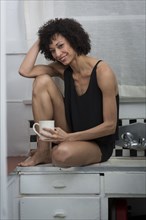 Mixed Race woman sitting on counter drinking coffee