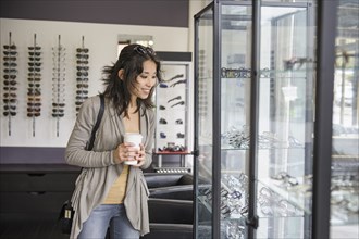 Mixed race woman drinking coffee and looking at eyeglasses in shop