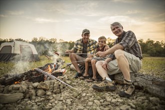 Three generations of Caucasian men roasting hot dogs over campfire