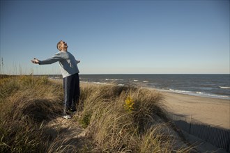 Man standing on sand dune near ocean with arms outstretched