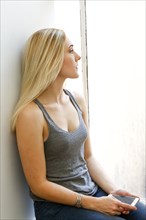 Pensive Caucasian woman sitting on windowsill holding cell phone