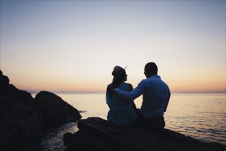 Silhouette of Caucasian couple sitting on rock near ocean at sunset