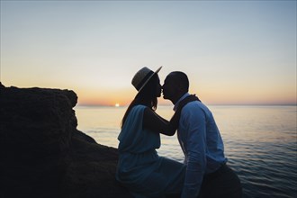 Caucasian couple kissing on rock near ocean at sunset