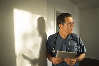 Hispanic doctor leaning on wall using digital tablet