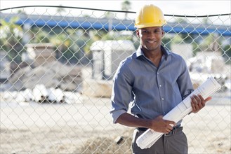 Black architect in hard hat holding blueprints near fence