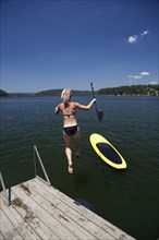 Caucasian woman jumping in lake next to paddleboard