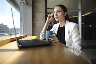 Pensive Caucasian woman drinking coffee near laptop