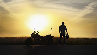Silhouette of man holding helmet near motorcycle at sunset