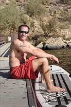 Hispanic man sitting on dock with paddleboard at river