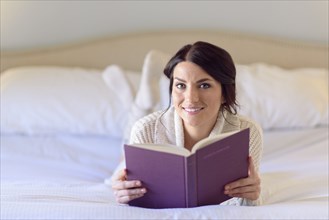 Smiling Caucasian woman laying on bed reading book