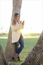 Hispanic woman leaning on tree texting on cell phone