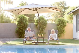 Caucasian couple reading newspaper and using laptop near swimming pool