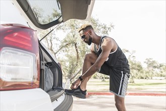 Black man leaning on car hatchback tying shoe