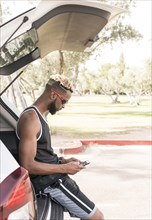 Black man leaning on car hatchback texting on cell phone