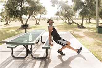 Black man leaning on picnic table in park stretching leg