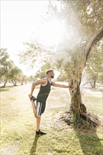 Black man leaning on tree in park stretching leg