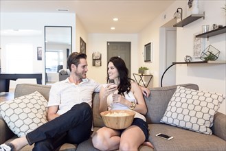 Smiling Caucasian couple relaxing on sofa with wine and popcorn