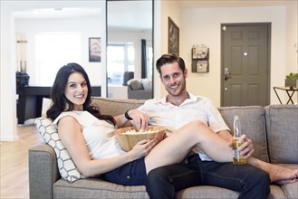 Smiling Caucasian couple relaxing on sofa with beer and popcorn