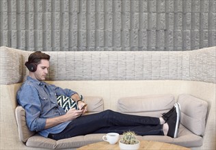 Caucasian man sitting on sofa wearing headphones texting on cell phone