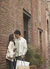 Young Asian couple leaning against a brick building kissing
