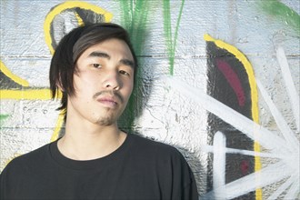 Young Asian man leaning against graffitied wall