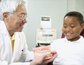 Senior Asian male dentist showing tooth model to young male patient
