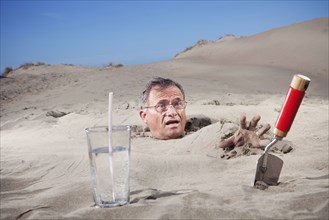 Caucasian man buried in sand next to glass of water and trowel
