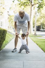 Father helping baby son walk on sidewalk